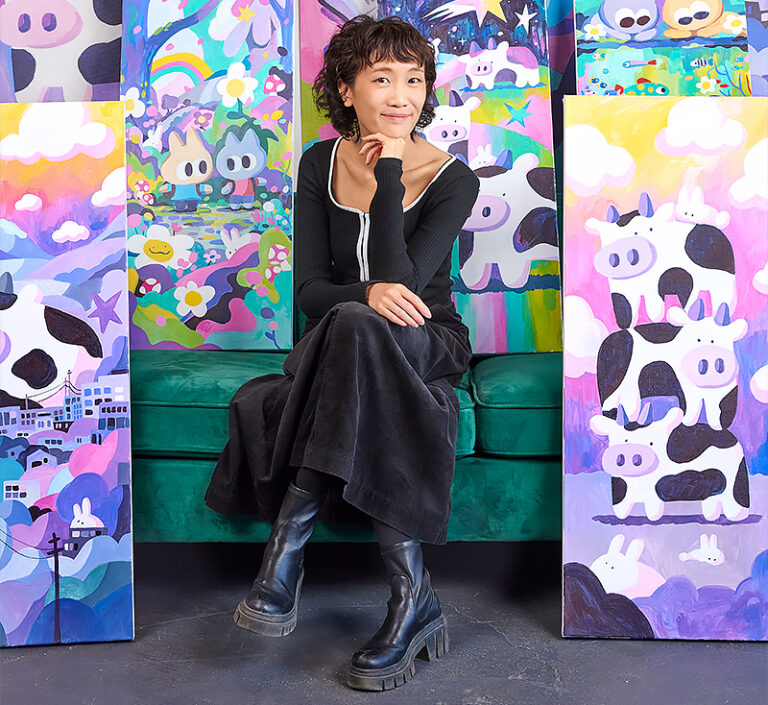 Artist Michelle Ku sits on a green velvet couch, surrounded by her vibrant, cartoon-style paintings featuring cows, rabbits, rainbows, and dreamy landscapes in pastel colours. She wears a black outfit and boots, smiling slightly at the camera.