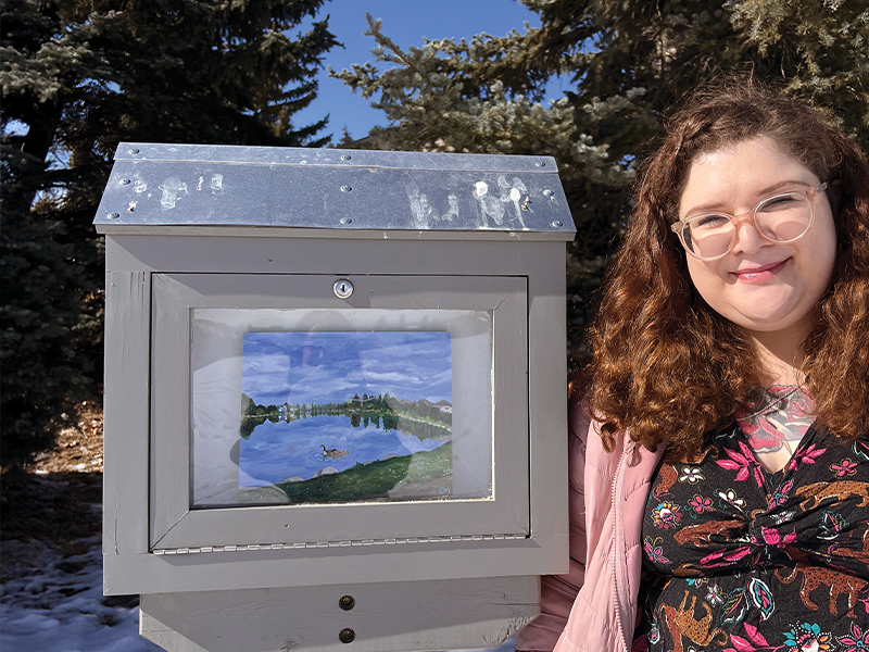 Erin McDonald next to her mini gallery showing her artwork titled "Coming Home" which seems to be a bright blue sky with forest and a duck on a pond that matches the sky's colour.