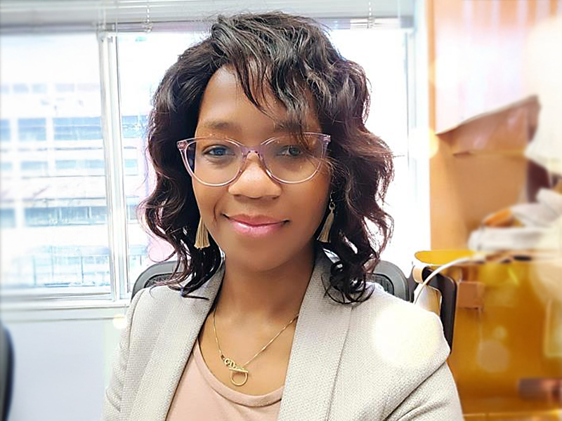 Gladys Marimira wearing glasses and a light grey suit jacket, black wavy hair smiling