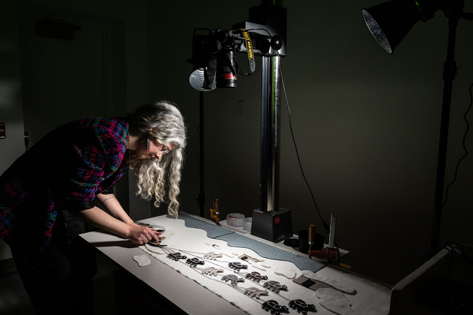 An artist assembles parts for what looks like an animation sequence using stop motion animation via an overhead camera. The long haired person with earrings is stooped over assembling the work.