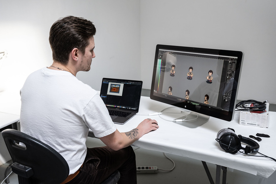 A student working on digital projects on a Mac laptop and large monitor. They have short hair and a forearm tattoo and nearby there is a pair of over-ear headphones.