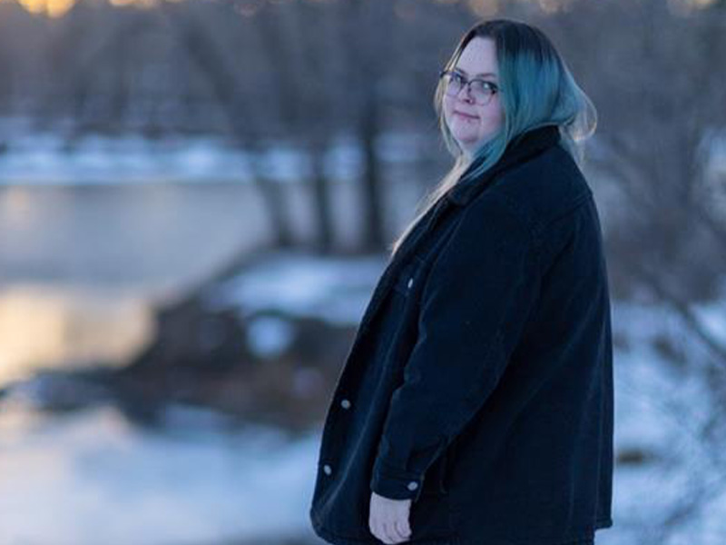 Jasmine Piper wearing a black coat standing in profile outside at sunset with snow on the ground and a blurred car in the background