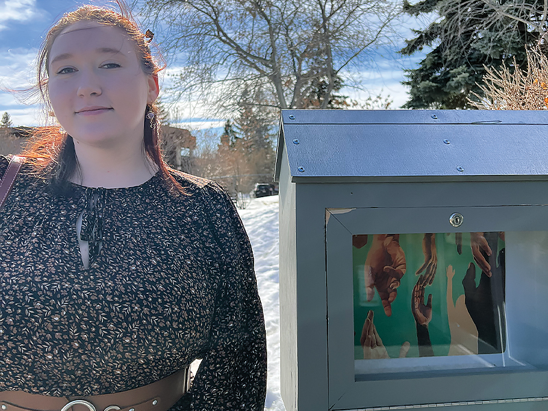 Artist Loriel Swoboda stands next to her mini gallery with artwork inside titled "Bond". Loriel is in a long sleeve shirt with brown leather belt tied around and some silver ornate features on the belt.