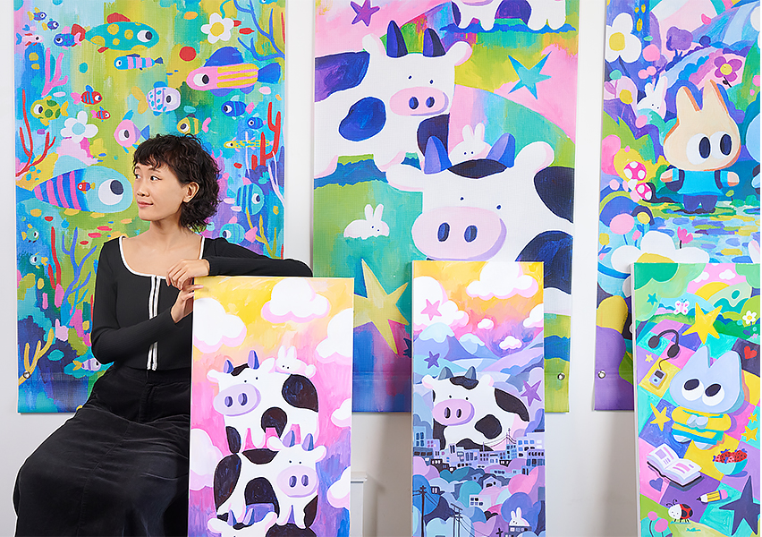 Artist Michelle Ku sitting on the left with banners she created next to her. The artwork is bright and colourful, almost cartoon like.