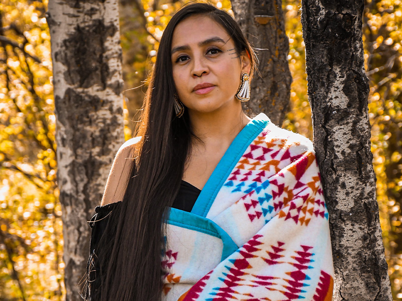 Riel Manywounds standing against birch trees in the forest with long dark hair down one side of her wrapped in an Indigenous wrap of white, blue, red and orange
