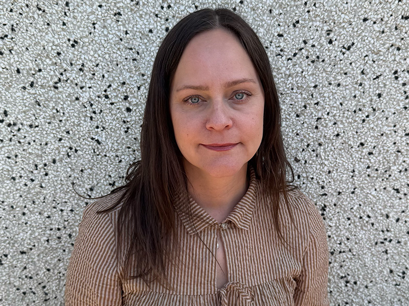 Sarah Houle with long dark hair wearing a brown buttoned shirt against a pebbled wall, smiling