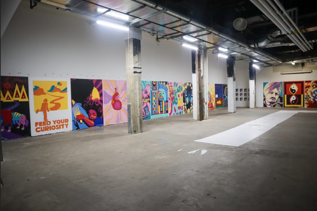 A large warehouse like studio, brightly lit with industrial ceiling, and large scale works of art lined up against the walls
