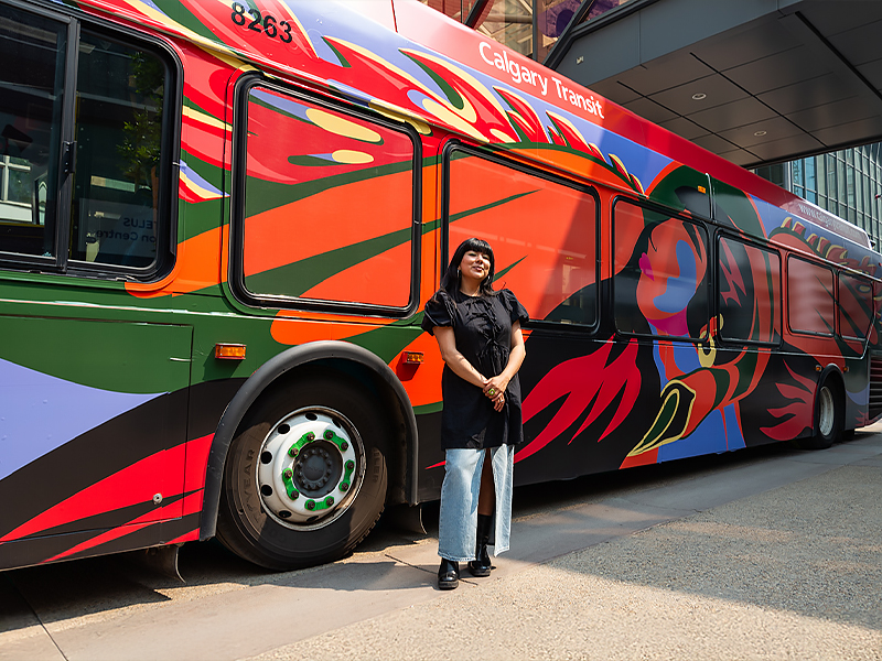 La Güita standing in front of a Calgary Transit bus with her Art Bus artwork covering it.