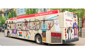 A Calgary Transit bus wrapped in a beige background, overlaid with colourful illustrations of Blackfoot dancers, horses, buffalo and symbolic patterns. The designs include Blackfoot words like iikakimaat and kimmaapiiyiptsin, along a pride flag. The bus is parked downtown, surrounded by brick buildings and trees.