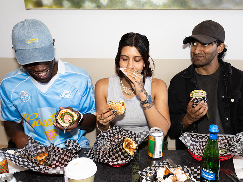 A man in a blue shirt and blue ballcap sits next to a woman in a grey tank top holding her hand to her mouth, with another man wearing black shirt and black ballcap on her other side, all eating wrapped sandwiches