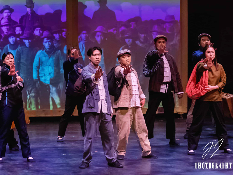 A group of performers on stage, dressed in traditional attire, stand in a powerful pose with one arm extended forward, palms open. They are mid-performance. Behind them is a projected image of historical black-and-white figures.