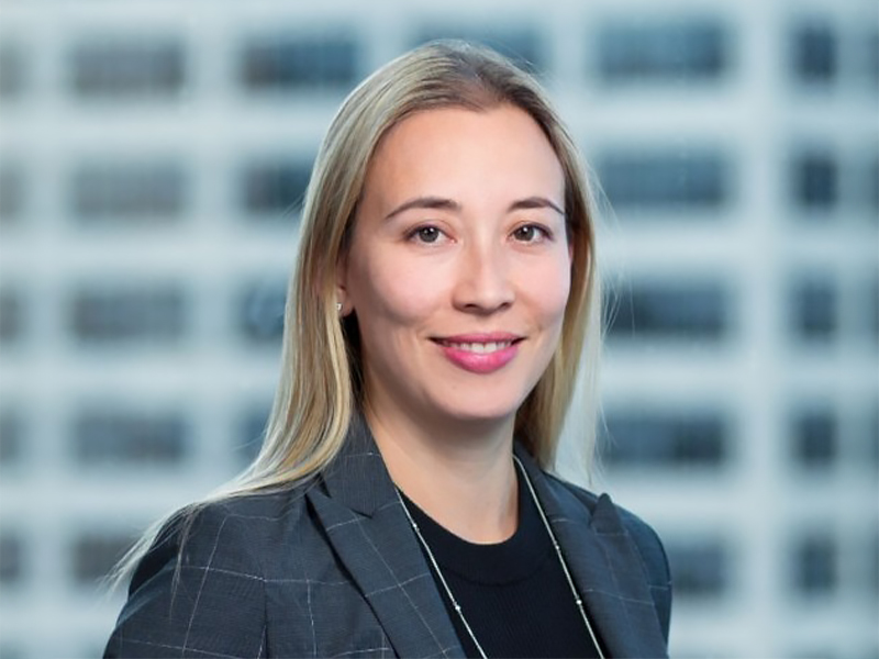 A headshot of Janet Huang wearing a dark blue suit jacket, black tshirt, long blond hair and smiling in front of a background that appears to be an office building