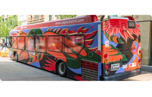 A Calgary Transit bus featuring bold red, orange and green wing-like shapes, with a womans face and a swan head abstractly painted on a dark base. It's parked beside stone buildings and a row of bright yellow chairs.