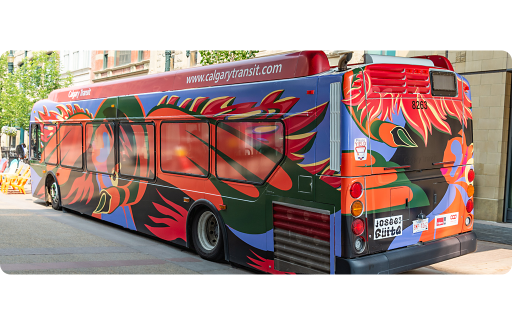 A Calgary Transit bus featuring bold red, orange and green wing-like shapes, with a womans face and a swan head abstractly painted on a dark base. It's parked beside stone buildings and a row of bright yellow chairs.