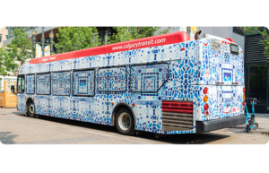 A Calgary Transit bus covered in a mosaic of blue and white tile patterns. It's parked along a downtown street near trees and scooters.