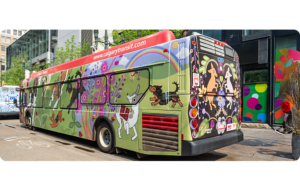 A Calgary Transit bus featuring colourful dogs, flowers and rainbows on a green and black background. Parked downtown surrounded by glass buildings and street art.
