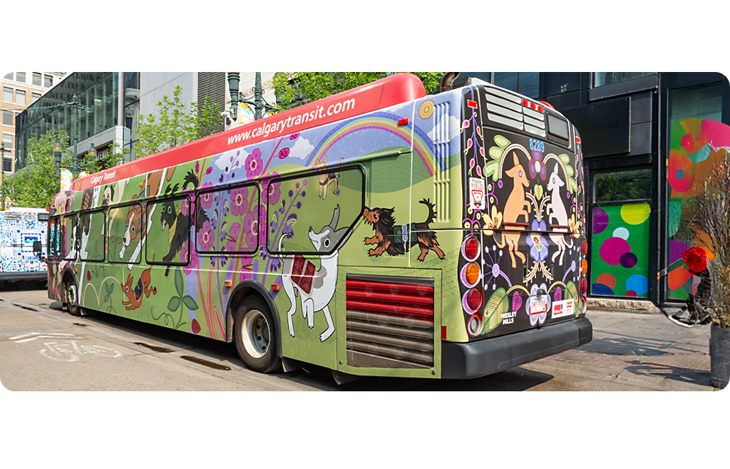 A Calgary Transit bus featuring colourful dogs, flowers and rainbows on a green and black background. Parked downtown surrounded by glass buildings and street art.