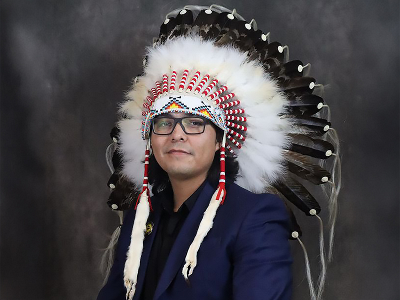 Richard Sparvier sitting against a dark background wearing an Indigenous headress and dark blue shirt with glasses.