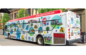 A Calgary Transit bus featuring a map-like illustration of Calgary. The background is white, with bold lines and solid colour circles in blue, yellow, red and green to highlight different sites. Landmarks like the Children's Hospital, City Hall and Peace Bridge, are shown in clear, simple illustrations alongside trees, pathways, and transit routes. The bus is outside and the background includes a downtown street with trees and buildings.