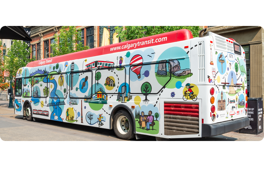 A Calgary Transit bus featuring a map-like illustration of Calgary. The background is white, with bold lines and solid colour circles in blue, yellow, red and green to highlight different sites. Landmarks like the Children's Hospital, City Hall and Peace Bridge, are shown in clear, simple illustrations alongside trees, pathways, and transit routes. The bus is outside and the background includes a downtown street with trees and buildings.