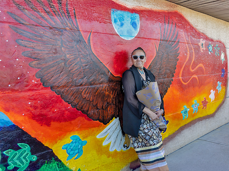 A woman in a dark shirt and long skirt stands against a mural of a red sky and eagle