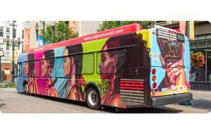 A Calgary Transit bus wrapped in bold artwork featuring side profiles of women in warm pinks, reds, blues and greens. The back shows a close-up of a person’s head and neck, hands holding the back of their own head. Their braids spell out “UNITY” in bold lettering. The bus is outside and the background includes a downtown street with banners and buildings.