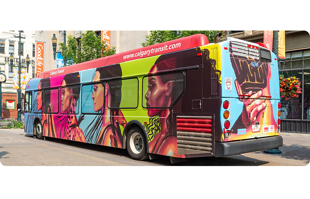 A Calgary Transit bus wrapped in bold artwork featuring side profiles of women in warm pinks, reds, blues and greens. The back shows a close-up of a person’s head and neck, hands holding the back of their own head. Their braids spell out “UNITY” in bold lettering. The bus is outside and the background includes a downtown street with banners and buildings.