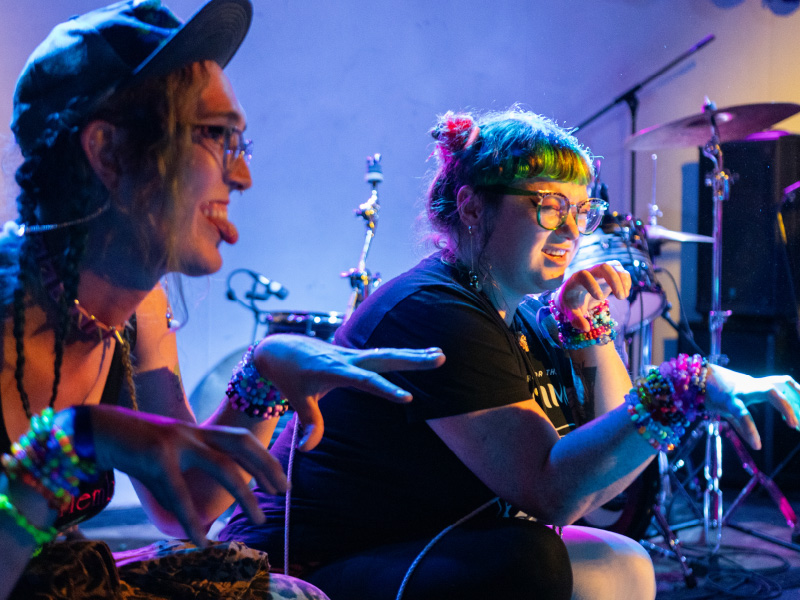 Two musicians perform with their hands raised to chest level, palm down, with fingers spread apart and pointed outwards. One is wearing a baseball cap, a spiked collar and is sticking their tongue out. The the other has coloured hair styled in space buns. Both are wearing glasses and a number of bracelets.