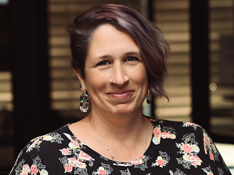 Jen Perrott smiling with a closed mouth wearing a black and flowered top, dark hair facing the camera