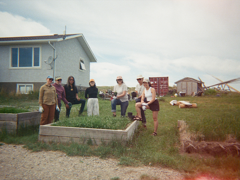 Six people pose in front of two raised garden beds outside a grey house. The group is smiling and wearing gardening gloves, hats and sunglasses. A red shipping container, a wooden shed and some tools are visible in the background