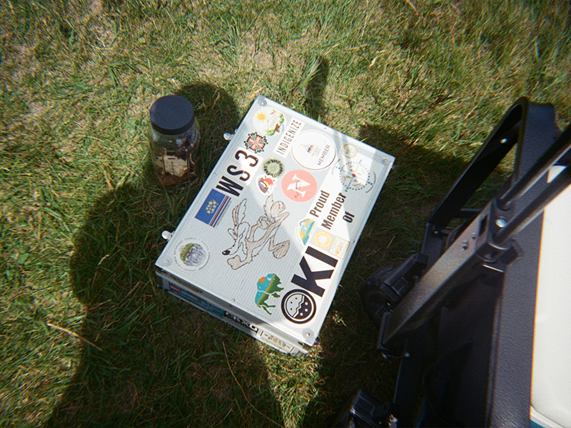 A small metal case covered in stickers sits on the grass. The stickers include text like “WS3,” “Proud Member of KEP,” and “Indigenize.” A drawing of a bird is in the centre. A black jar is beside the case, and the wheel of a cart is partly visible on the right.