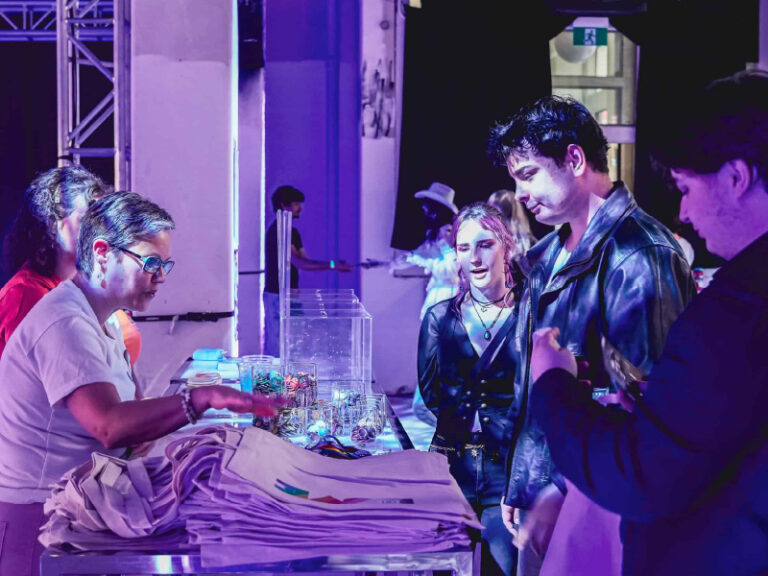 Participants at an Alphabet Mafia Collective event gather around a display table. The room is bathed in a purple light.