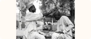 A man gives another man a haircut outdoors. Both are seated on a wooden bench. The barber holds a razor while the other man leans forward slightly, eyes closed. The photo is in black and white.