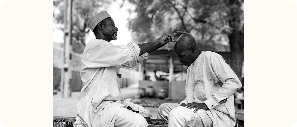 A man gives another man a haircut outdoors. Both are seated on a wooden bench. The barber holds a razor while the other man leans forward slightly, eyes closed. The photo is in black and white.