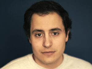 Rino Mioc from the shoulders up looking forward, wearing a light coloured shirt against a dark background