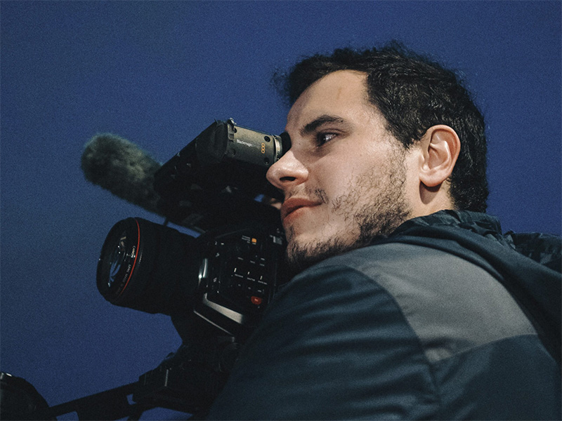 Rino Mioc wearing dark clothes holding a video recorder up as if shooting a scene for a film