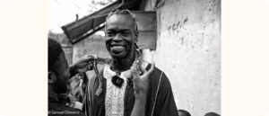A black and white photo of a smiling man stands outdoors while another person places a patterned snake around his shoulders. The man wears traditional clothing with decorative embroidery.