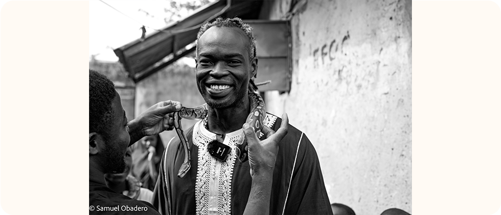 A black and white photo of a smiling man stands outdoors while another person places a patterned snake around his shoulders. The man wears traditional clothing with decorative embroidery.