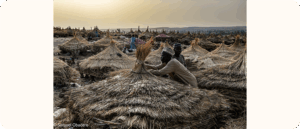 Two men crouch beside a cluster of round thatched roofs in an open field at sunset, surrounded by many similar structures.