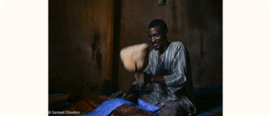 A Black man sits on the floor inside a dimly lit room, smiling as he pounds blue fabric with a wooden tool.