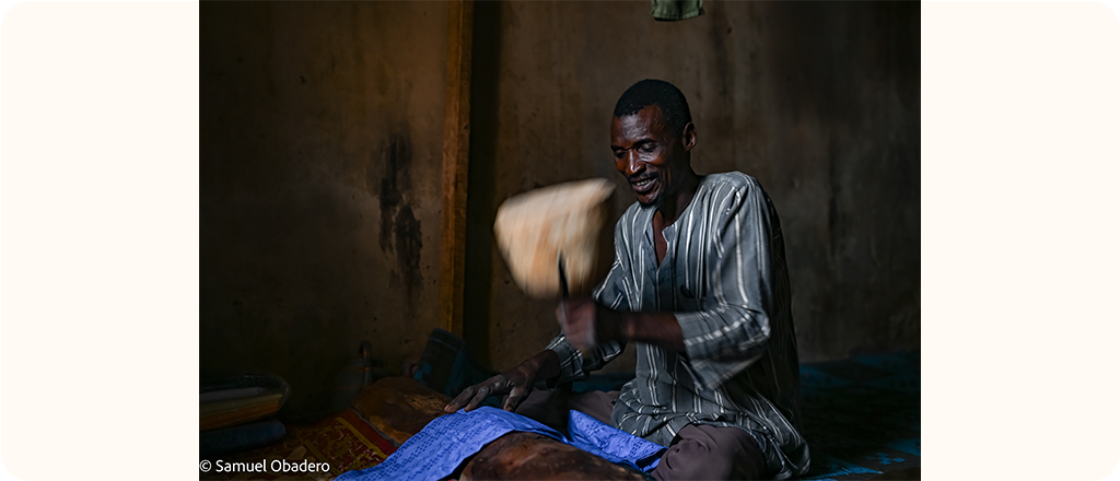 A Black man sits on the floor inside a dimly lit room, smiling as he pounds blue fabric with a wooden tool.