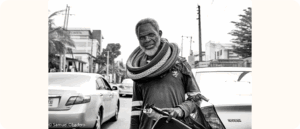 Black and white photograph of an older man standing in the street between cars. He is carrying car parts and has several tires around his shoulders and neck. Buildings, cars and utility poles are visible in the background.