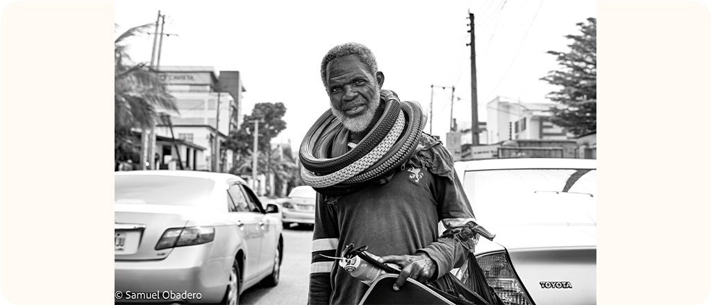 Black and white photograph of an older man standing in the street between cars. He is carrying car parts and has several tires around his shoulders and neck. Buildings, cars and utility poles are visible in the background.