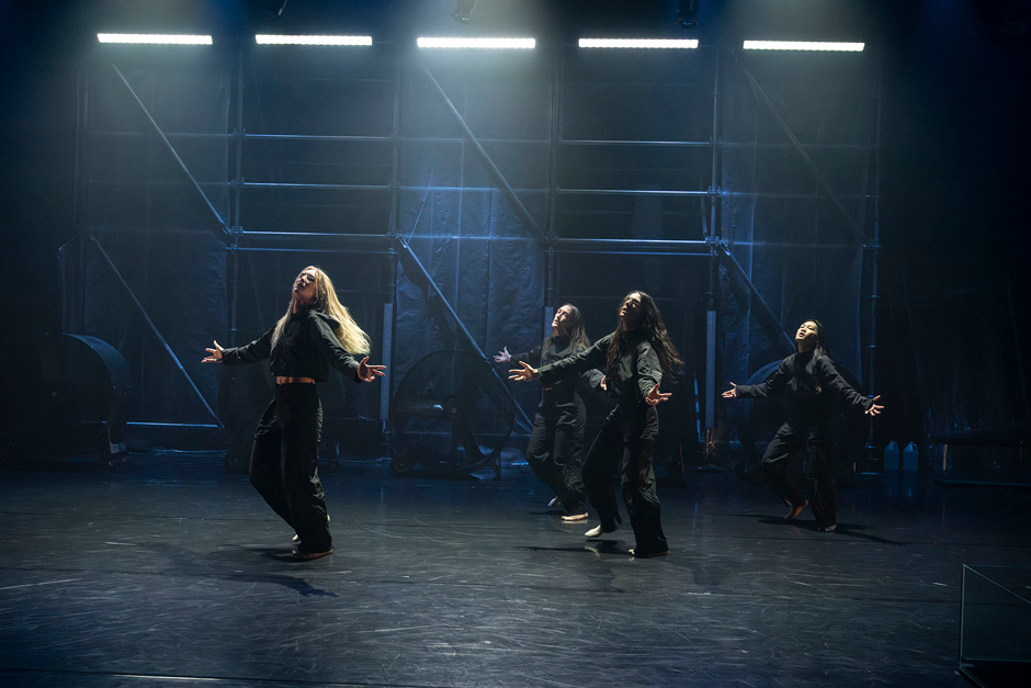 Dance performers on a stage or a set singing and performing. The performers feel like they are all mixed gender and racial background. they all seem to have long hair, and dark costumes.