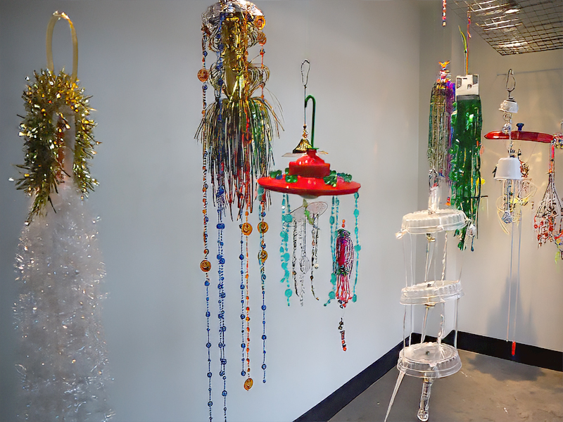 Installation view of a corner gallery with multiple vibrant, handcrafted hanging sculptures made from beads, plastic, tinsel and fringe. The ornaments are hanging from the ceiling, casting soft shadows on the white walls.