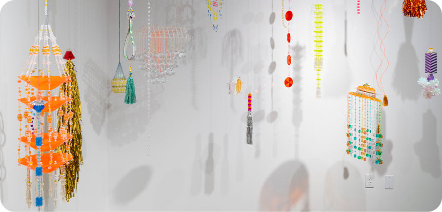 A group of hanging sculptures made from colourful beads, tassels, and found materials suspended from the ceiling in a white gallery space. The pieces include bright orange trays, gold fringe, neon yellow rods, and transparent elements that cast soft shadows on the wall.