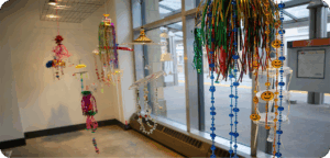 A group of colourful, handmade hanging sculptures made from beads, bells, plastic trinkets and ribbons. The decorations dangle from the ceiling in front of a window.