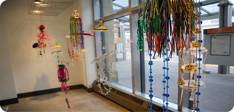 A group of colourful, handmade hanging sculptures made from beads, bells, plastic trinkets and ribbons. The decorations dangle from the ceiling in front of a window.