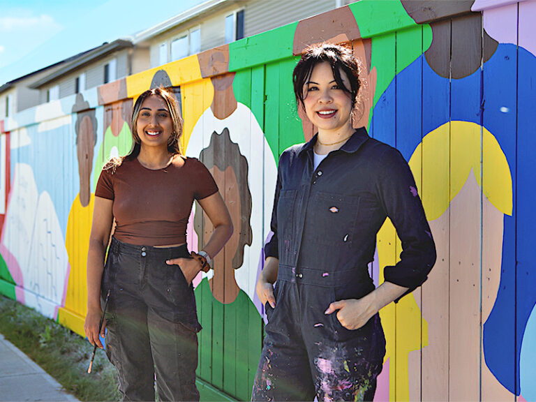 Two artists stand smiling in front of a brightly coloured mural painted on a wooden fence. The artwork features large, figures in pink, green, blue, yellow and brown. Artist on the left wears a brown shirt and black overalls; the artist on the right wears a dark navy jumpsuit with paint splatters.