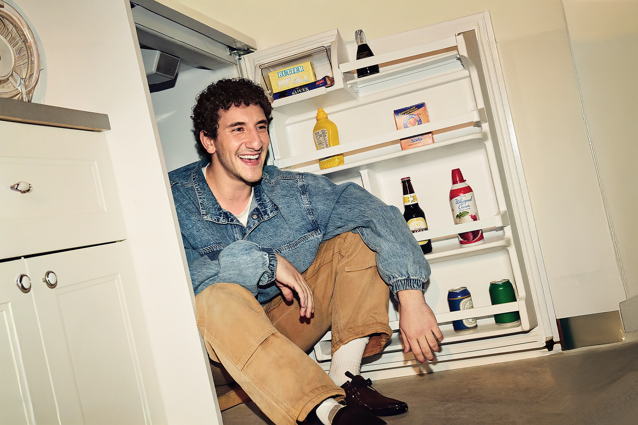 Malik Elassal sitting with a broad smile, within a small refrigerator which has bottles in the doorway, in a kitchen